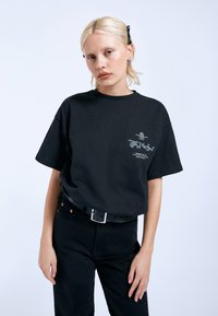 Černé bavlněné tričko s krátkým rukávem a grafickým potiskem na hrudi. Model ho nosí s černými kalhotami a černým pásem.