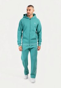 Turquoise zip-up hoodie met een tweekleurig ontwerp, voorzien van een zak aan de voorkant en witte strepen op de broek. Gemaakt van een zachte, gestructureerde stof.