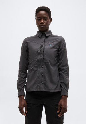 Person trägt ein dunkelgraues Hemd mit langen Ärmeln und Knöpfen, das ein dezentes Logo auf der Brust hat, und schwarze Hosen, steht frontal vor einem schlichten Hintergrund.