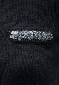 Črno pleteno blago na predelu prsnega žepa vsebuje horizontalni vrstico sijočih srebrnih sequinov, ki zagotavlja kontrast teksture in detajle.