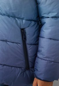 Kurtka ocieplana w gradientowych odcieniach niebieskiego, z boczną kieszenią na suwak, gładką teksturą i pikowanymi sekcjami dla lepszej izolacji.