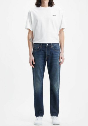 Personne portant un t-shirt blanc Levi's, un jean bleu foncé, des baskets blanches, debout devant un fond blanc uni, les bras détendus.