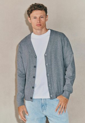 Young man with curly hair wearing a gray buttoned cardigan over a white t-shirt and light blue jeans, standing against a plain background.