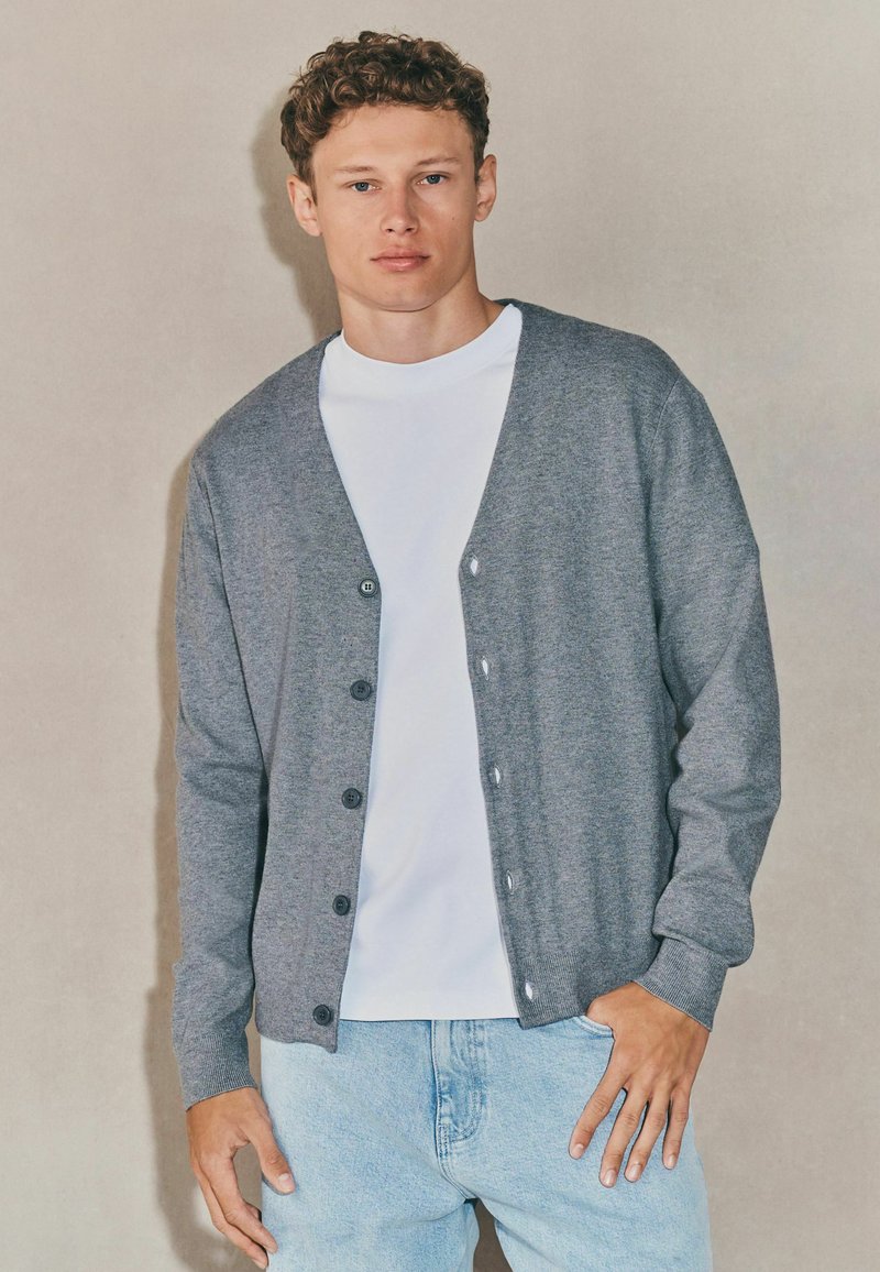 Tânăr cu păr creț, îmbrăcat cu un cardigan gri cu nasturi peste un tricou alb și blugi deschiși, stând în fața unui fundal simplu.