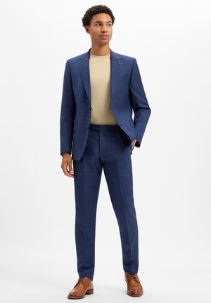Man standing wearing a blue suit with a beige shirt and brown dress shoes against a plain white background.