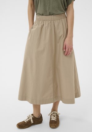 Donna che indossa una gonna midi beige con fascia elastica in vita e sneakers marroni con lacci bianchi, in piedi davanti a uno sfondo bianco uniforme.