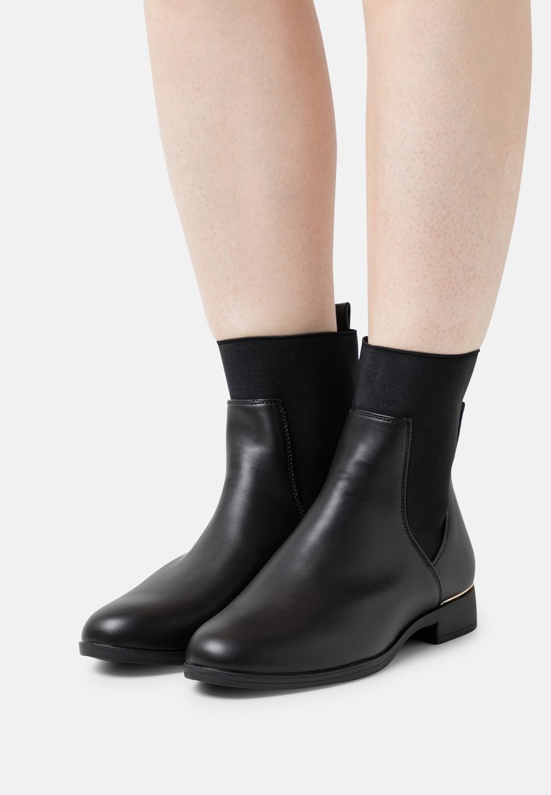 Jambes portant des bottines noires avec des parties avant en cuir lisse et des parties supérieures en tissu extensible, debout sur une surface blanche.