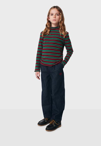 Raidallinen pitkähiha-paita punaisessa ja vihreässä, yhdistetty tummansinisiin, ohuilla raidoilla kuvioituihin housuihin, joissa on punainen logo. Mustat kengät, joissa ruskeat pohjat.