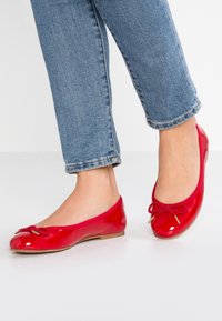 Person med blå jeans och glansiga röda ballerinasandaler med rosetter, stående på en vit golv.