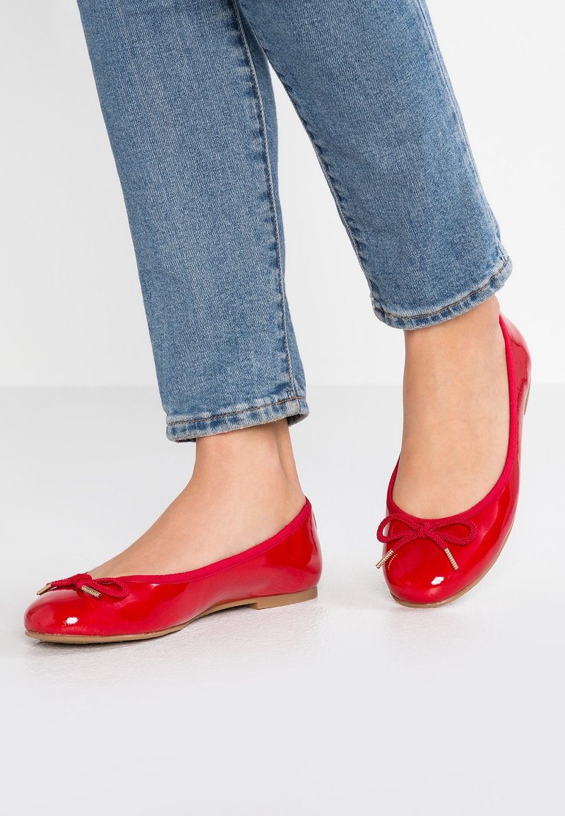 Person med blå jeans och glansiga röda ballerinasandaler med rosetter, stående på en vit golv.