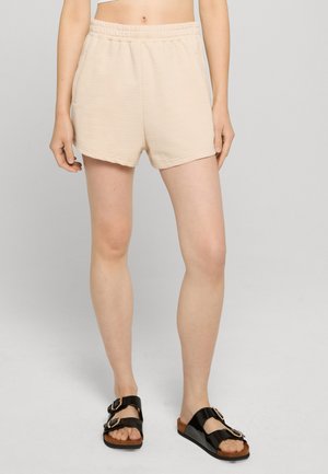 Personne portant un short beige côtelé à taille élastique et des sandales noires à double boucle, debout devant un fond gris clair.