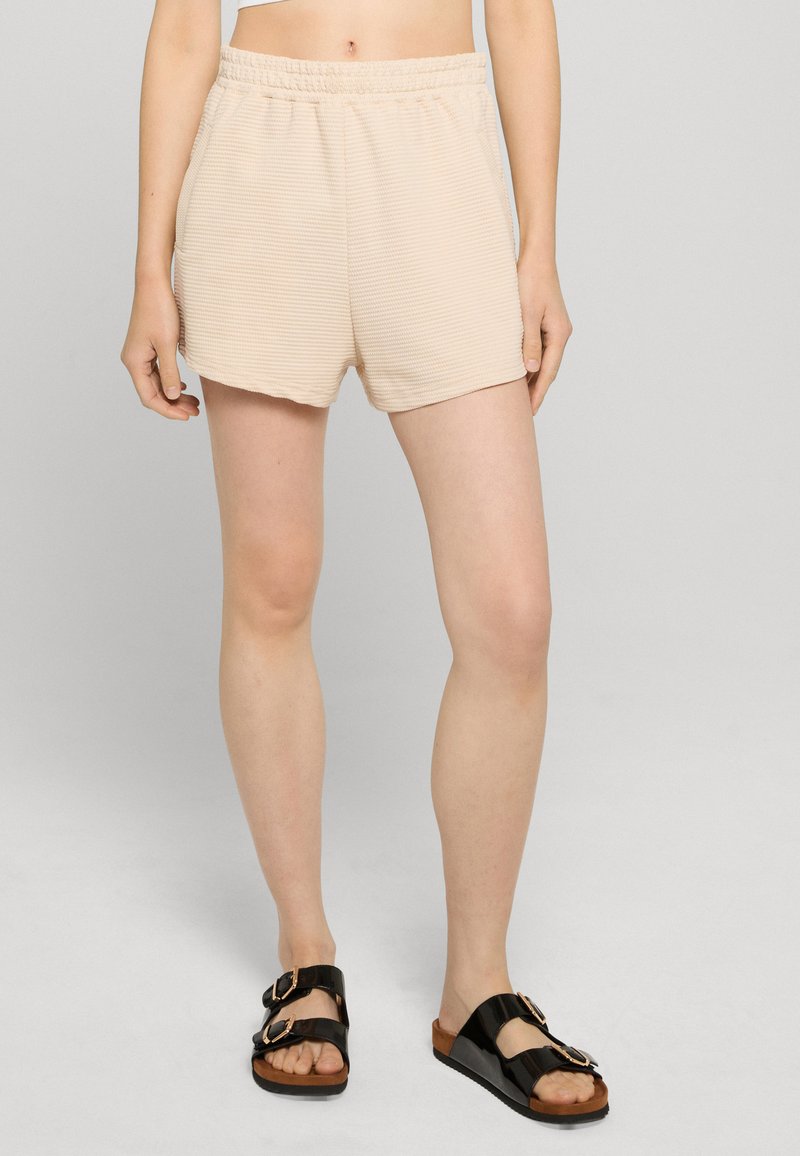 Personne portant un short beige côtelé à taille élastique et des sandales noires à double boucle, debout devant un fond gris clair.