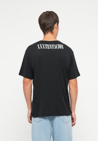 Sort bomulds T-shirt med teksten "XXXTENTACION" i hvid på den øverste del af ryggen, korte ærmer og en afslappet pasform.