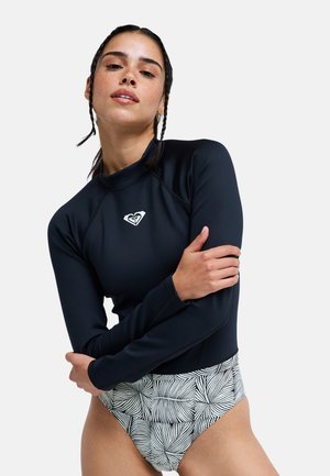 Kobieta z plecionymi włosami nosząca czarną koszulkę rash guard z długim rękawem z białym logo i czarno-białe wzorzyste majtki bikini.