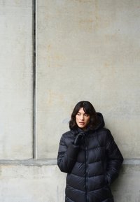 Čierna prešívaná bunda s kapucňou, so zipsovým uzáverom. Hladká textúra, stredná dĺžka, s rukavicami a stojaca pri betónovej stene.