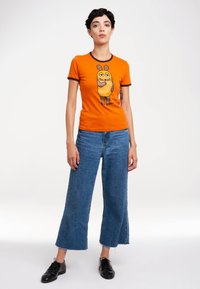Orange T-shirt with brown-trimmed neckline and sleeves, featuring a cartoon character graphic. Paired with blue wide-leg jeans and black shoes.