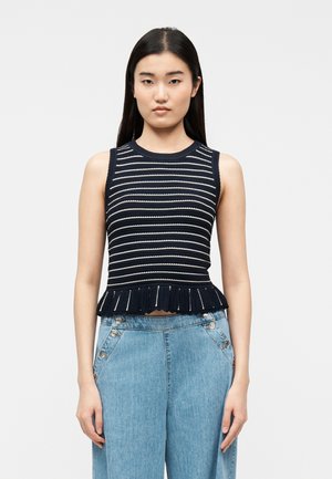 Ung kvinne med langt mørkt hår iført en ermeløs marineblå og hvitstripet strikket topp med volangkant og høytlivs blå jeans.