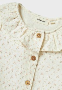 Camicia bottonata color crema con un delicato motivo floreale. Presenta un colletto arricciato, colletto a volant e bottoni in legno.