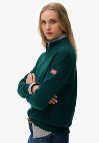 Donkergroene ritsvest met hoge kraag, geribbelde manchetten en zoom. Bevat een decoratieve patch op de mouw en een gelaagd gestreept shirt eronder.