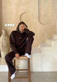 Brun huvtröja och set med sweatpants med en logo. Sitter på en träpall, vita strumpor, i en mjukt texturerad, neutral-tonad inredning.