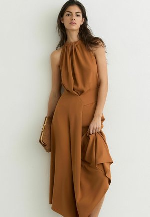 Kobieta w bezrękawnej, ceglano-pomarańczowej sukience z odkrytymi ramionami, trzymająca pasującą kopertówkę i tkaninę, stojąca na tle białej ściany.