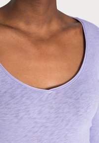 Jasnofioletowa bluzka krótki rękaw z głębokim dekoltem w kształcie litery V i miękkim, teksturowanym materiałem. Dekolt zdobi subtelny, kontrastowy wykończenie.