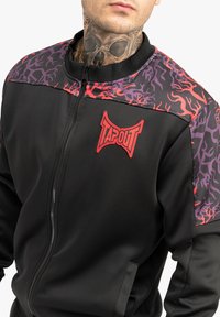 Crna jakna na zip s apstraktnim uzorkom u ljubičastoj i crvenoj boji na ramenima. Ima crveni "TAPOUT" logo na prsima i glatku teksturu.