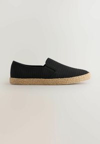 Zapato negro texturizado sin cordones con suela trenzada de yute color canela, perfil bajo y detalles elásticos en los lados sobre un fondo neutro.