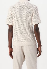 Suéter beige de punto con mangas cortas, con un patrón texturizado y dobladillo acanalado. La parte trasera tiene un diseño transpirable con aberturas verticales.