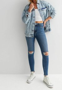 Kvinna iklädd ljus denimjacka, vit kort t-shirt, trasiga skinny jeans och vita högklackade sneakers, stående mot en enkel vägg.