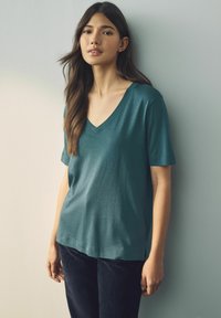 T-shirt teal à encolure V en tissu léger avec des manches courtes, coupe décontractée et fentes sur les côtés. Associé à un pantalon foncé sur un fond neutre.
