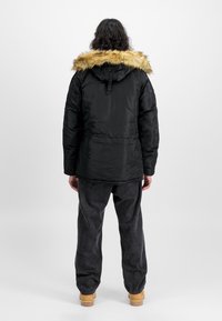 Parka aislante negra con capucha forrada de piel, que presenta una textura suave y un ajuste ligeramente ceñido en la cintura, usada sobre pantalones vaqueros oscuros.