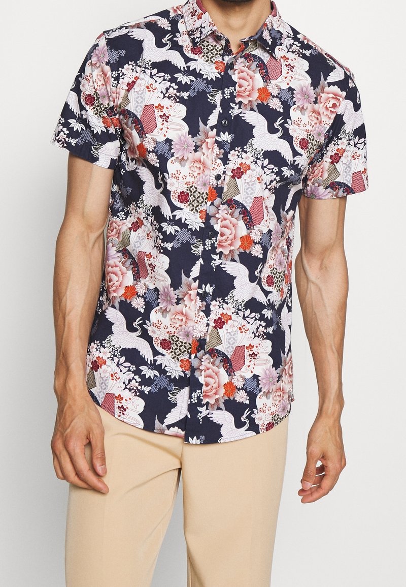 Chemise homme à manches courtes boutonnée, fond bleu marine, imprimé floral et grue, portée avec un pantalon beige.