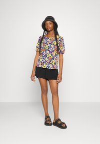 Top de mangas curtas com padrão floral sobre um fundo azul-marinho, calções plissados pretos e sandálias pretas com studs e fivelas. Textura suave por todo o conjunto.