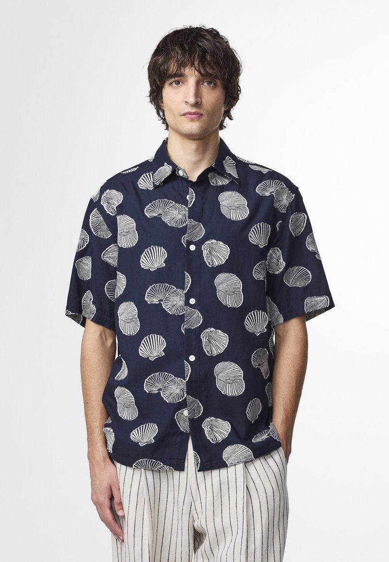Camicia navy a maniche corte con motivi a conchiglia bianchi, bottoni frontali e colletto. Abbinata a pantaloni a righe di colore chiaro.