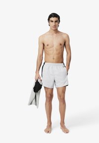 Lichtgrijze zwembroek met zwarte zijaccenten, elastische tailleband en een klein logo. Model houdt een grijze handdoek en zwart shirt vast, op blote voeten.