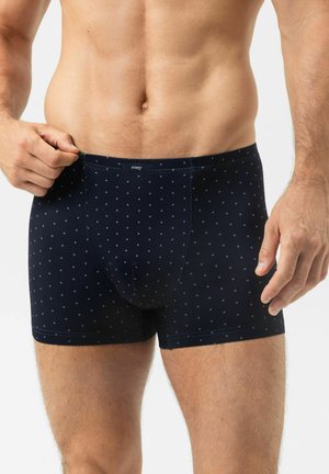 Mann trägt marineblaue Boxer-Shorts mit kleinen hellblauen Punkten und richtet den Bund mit der Hand auf weißem Hintergrund.