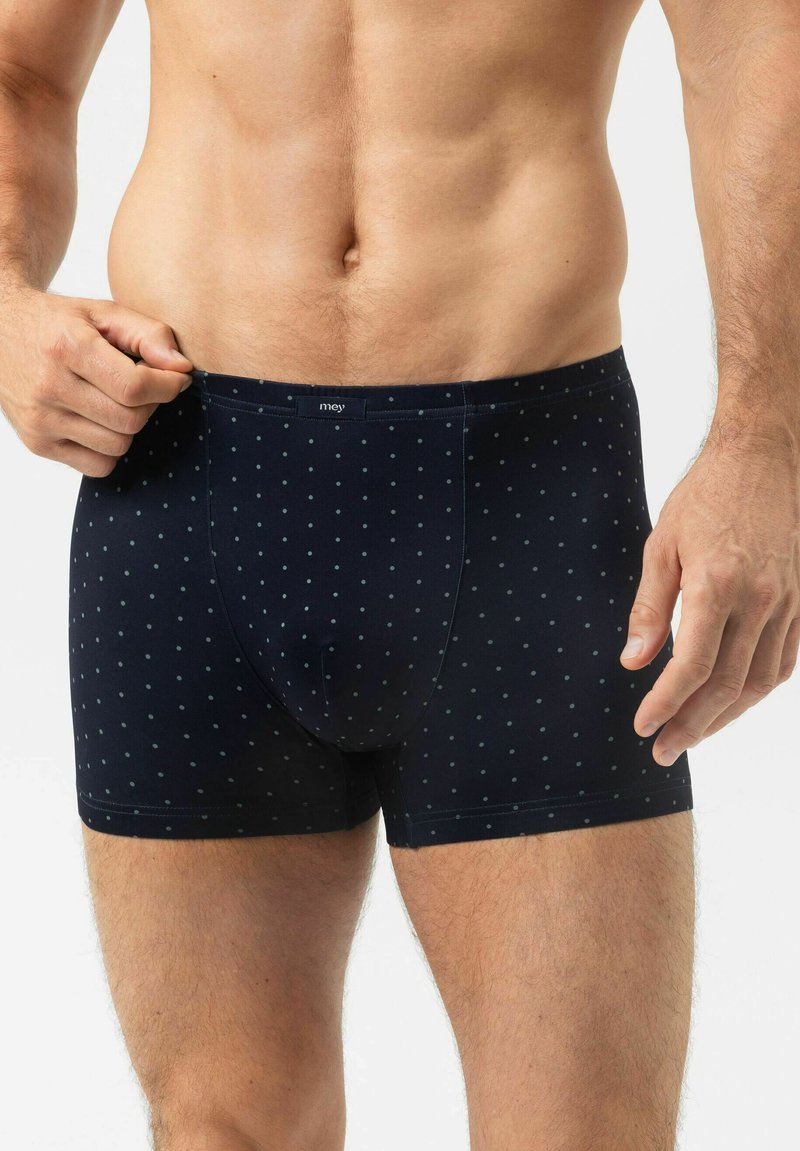 Man wearing navy blue boxer briefs with small light blue polka dots, adjusting waistband with hand on a white background.