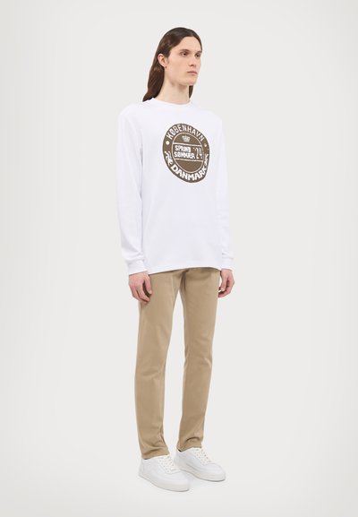 Long-sleeve white T-shirt with a round brown graphic design, paired with beige pants and white sneakers, standing on a light background.