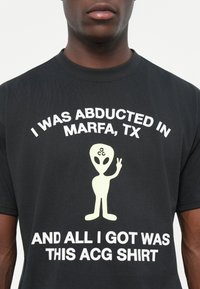 Svart bomull t-shirt med vit text som säger "JAG BLEV ABDUKTAD I MARFA, TX OCH ALLT JAG FICK VAR DENNA ACG TRÖJA" med en alien grafik.