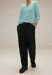 Hellblauer, gestrickter Pullover mit V-Ausschnitt, kombiniert mit schwarzen, weiten Hosen. Der Pullover hat gerippte Bündchen; die Hosen verfügen über Fronttaschen und einen Knopf.