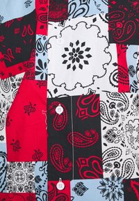 Camicia patchwork con una combinazione di motivi paisley e floreali in rosso, nero, bianco e blu, dotata di due bottoni bianchi e realizzata in cotone morbido.