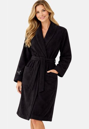 Frau trägt einen schwarzen Fleece-Bademantel mit Schalkragen, Gürtel, Vordertaschen und Puma-Logo am linken Ärmel, lächelt in die Kamera.