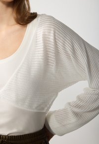 Vit topp med ett andningsbart meshdesign med horisontella ränder, långa ärmar och vid halsringning, lager-på-lager över ett enfärgat baslager.