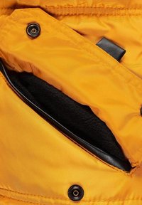 Veste rembourrée jaune avec une poche doublée en polaire noir, mettant en valeur un accent en cuir noir et une fermeture à bouton-pression.