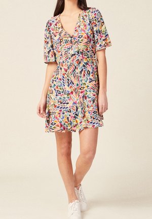 Femme portant une robe portefeuille courte à motifs colorés avec des manches courtes et des baskets blanches, debout devant un fond beige uni.