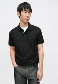 Polo nera con motivo a rombi strutturato, maniche corte e colletto. Indossata con jeans grigio scuro. Semplice abbottonatura sul collo.