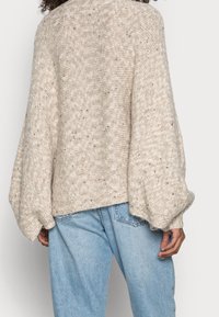 Beiger übergroßer Strickpullover mit Ballonärmeln, mit einer strukturierten Oberfläche und subtilen Sprenkeln, kombiniert mit hellblauen Jeans.