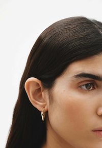 Pendientes en tono dorado con forma de punta de flecha, colgando de un pequeño aro, exhibidos en la oreja de un modelo. Superficie lisa y pulida.