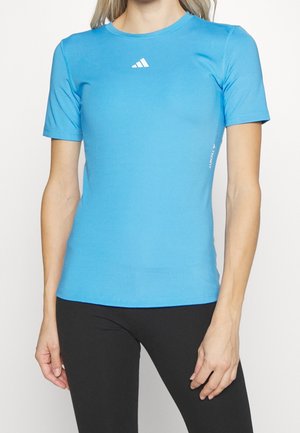 Kobieta nosząca dopasowaną jasnoniebieską sportową koszulkę Adidas z krótkim rękawem i czarne legginsy, pokazana od ramion do bioder.